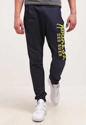 Homme portant un jogging bleu marine foncé avec le texte jaune « BOXEUR DES RUES » sur la cuisse gauche et des baskets blanches, marchant vers l'avant sur un fond uni.