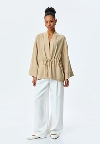 Beige offener Kimono mit übergroßen Ärmeln, cinchbarem Bund und zwei Fronttaschen, kombiniert mit weiten weißen Hosen und spitzen weißen Schuhen.