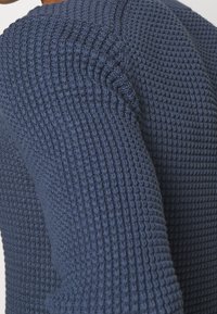 Gros plan sur la manche d'un pull tricoté bleu marine texturé avec un motif en nid d'abeille, montrant les détails des coutures et la texture du tissu.