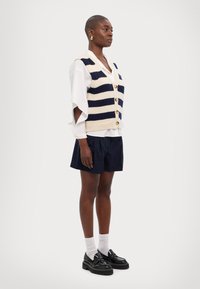 Triibuline kudumivest kreemjas ja tumesinis toonis, V-kaelusega ja nööpidega eest. Ühendatud valge ülemõõdulise särgi ja tumesiniste lühikeste pükstega. Mustad sarvajalanõud.