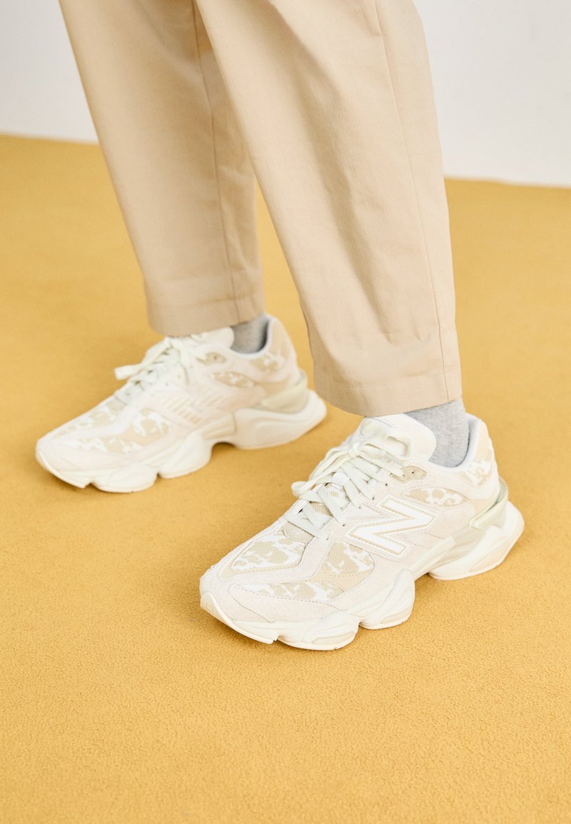 Zapatillas deportivas blancas con un upper texturizado que presenta patrones sutiles, forma redondeada y suela acolchada, combinadas con pantalones beige.