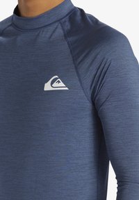 Tamno plava dugak rukav zaštita od sunca s visokim ovratnikom, napravljena od glatkog, rastezljivog materijala i s bijelim logotipom na prsima.