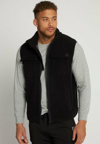Schwarze Fleece-Weste mit Reißverschluss, zwei Brusttaschen, Seitentaschen und hohem Kragen. Getragen über einem grauen Langarmshirt.
