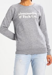 Grijze sweatshirt met geribde manchetten en zoom, met witte tekst "Abercrombie & Fitch Co." over de voorkant. Draagt lichtblauwe jeans met slijtageplekken.