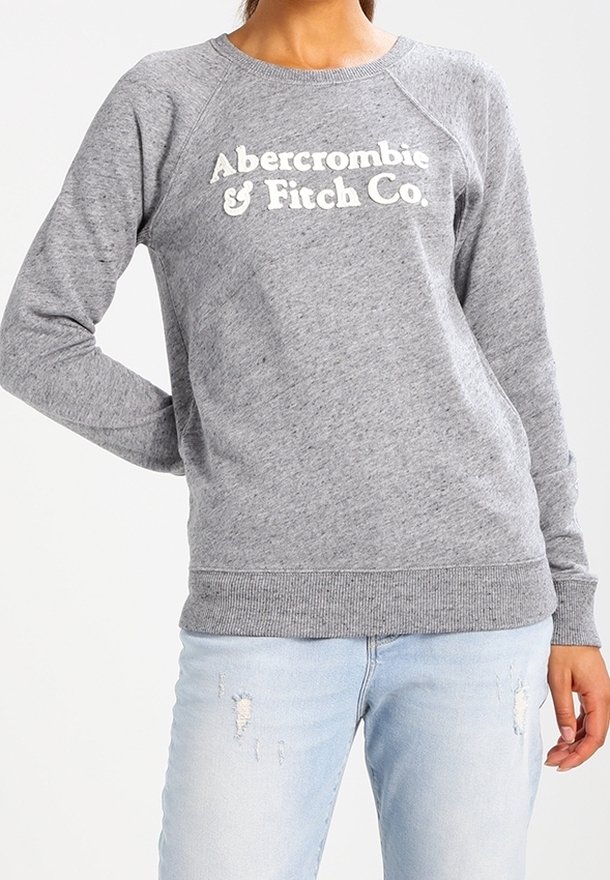 Grijze sweatshirt met geribde manchetten en zoom, met witte tekst "Abercrombie & Fitch Co." over de voorkant. Draagt lichtblauwe jeans met slijtageplekken.