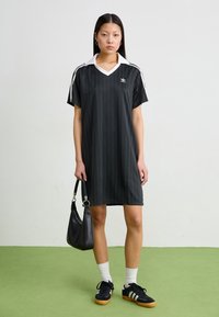 Woman stands on green floor wearing black Adidas dress with white collar, black sneakers, white socks, and holding a small black handbag.