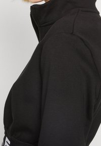 Svart zippered jacka med hög krage, tillverkad av ett slätt tyg. Har en bekväm passform och en subtil logotyp längs fållen.