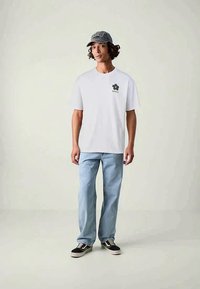 Λευκό βαμβακερό μπλουζάκι με μαύρη γραφική απεικόνιση λουλουδιού στο αριστερό στήθος, συνδυασμένο με ανοιχτού μπλε τζιν παντελόνι και μαύρα αθλητικά παπούτσια.