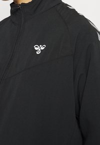 Chaqueta negra con cremallera y cuello alto, textura suave y un pequeño logo blanco en el pecho; acentos a rayas en las mangas.