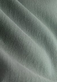 Țesătură moale și texturată în verde mat, cu nervuri diagonale și cute netede, indicând un material textil ușor.