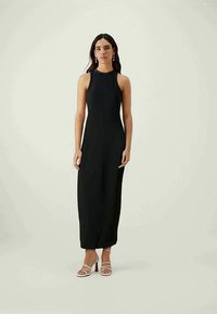 Melna bezpiedurkņu maksi kleita ar augstu kakla izgriezumu, pieguļošu siluetu un gludu tekstūru, papildināta ar striķa gaismas toņa sandālēm.