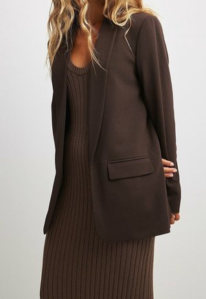 Femme portant une longue robe en maille côtelée marron foncé et un blazer assorti ajusté avec une poche à rabat, posant devant un fond clair uni.