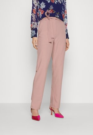 Kvinde iført lyserøde bukser med bindebånd i taljen, blomstret marineblå bluse og lyserøde højhælede slingback-sko, stående mod hvid baggrund.