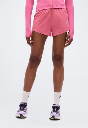 Frau trägt pinke Sportshorts, ein pinkes Langarmshirt, weiße Knöchelsocken und lila Laufschuhe, steht vor einem schlichten Hintergrund.