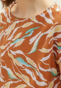 Frau, die eine lockere orangefarbene Bluse mit abstrakten weißen, gelben und türkisfarbenen blattartigen Mustern und einem runden Ausschnitt trägt.