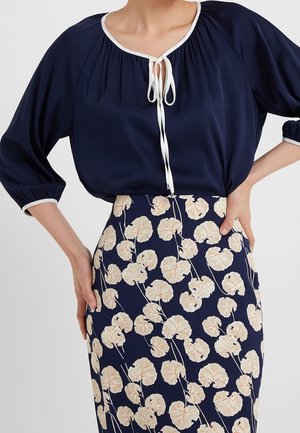 Granatowa bluzka z białym obszyciem i wiązaniem przy dekolcie, zestawiona z granatową spódnicą z wysokim stanem, ozdobioną beżowym wzorem kwiatowo-liściastym.
