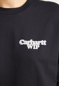 Melna kokvilnas T-krekls ar baltu izšuvumu "Carhartt WIP" logotipu kreisajā krūšu daļā. Apaļa kakla forma, īsas piedurknes.