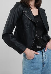 Giacca da moto nera in pelle con chiusura a zip, hardware argentato e design aderente, abbinata a un top nero con paillettes e jeans azzurri chiari.