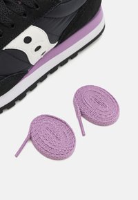 Sneaker nera con accenti grigi e bianchi, dotata di una suola intermedia viola. Due stringhe rotonde lavanda posizionate accanto al sneaker.