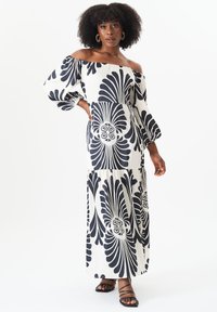 Maxi dress with off-shoulder neckline, black and white floral print, puff sleeves, and tiered design. Finished with thin straps on sandals.