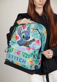 Mochila con un personaje de dibujos animados, predominantemente de color azul claro con patrones florales, y la palabra "STITCH" en negrita. Incluye múltiples bolsillos con cremallera.