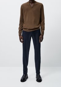 Homme portant un pull marron à col en V, un pantalon bleu marine foncé avec des revers, et des bottes en cuir noir, debout devant un fond clair uni.