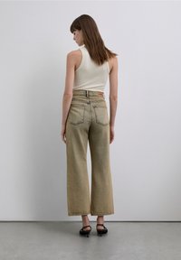 Jeans beige chiaro a gamba larga con cinque tasche, cuciture visibili e una targhetta con marchio. Il modello indossa una canotta bianca aderente e tacchi neri.