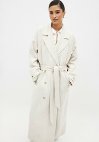 Cappotto lungo beige realizzato in tessuto testurizzato, con chiusura doppiopetto, ampie rever e una cintura legata in vita.