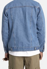 Denimjack in lichtblauw, met een klassieke snit, contrasterende stiksels en metalen hardware. Gelaagd over een witte blouse en kaki broek.