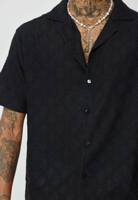 Chemise noire à manches courtes avec un motif en losanges texturé et un col. Présente des boutons noirs et est portée sur une peau tatouée.