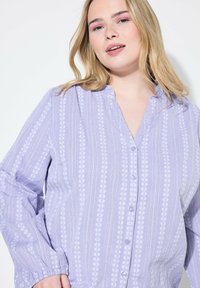Camisa de botones de color púrpura claro con una suave textura, que presenta un sutil patrón vertical de motivos florales blancos y puños fruncidos.
