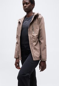 Άτομο που φοράει μια μπεζ κουκούλα Columbia πάνω από μαύρο πουκάμισο Salomon και μαύρο παντελόνι, στέκεται μπροστά σε μια απλή ανοικτή φωτεινή επιφάνεια.