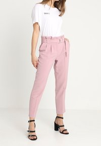 Pantalons ajustés rose clair avec une taille ceinturée, des poches avant et une coupe slim, associés à un t-shirt blanc avec une inscription noire.