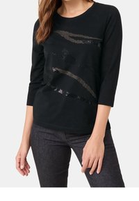 Donna che indossa una maglietta nera a maniche lunghe con motivi astratti in paillettes, abbinata a jeans aderenti grigio scuro, in piedi davanti a uno sfondo neutro.