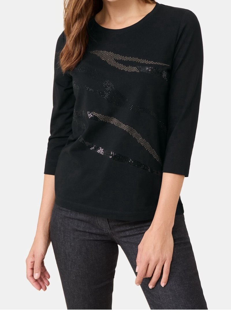Donna che indossa una maglietta nera a maniche lunghe con motivi astratti in paillettes, abbinata a jeans aderenti grigio scuro, in piedi davanti a uno sfondo neutro.