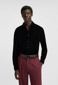 Schwarzes Samt-Hemd mit langen Ärmeln, Kragen und Knöpfen, kombiniert mit hoch taillierter bordeauxroter Hose und schwarzem Ledergürtel.