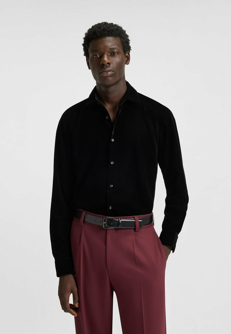 Schwarzes Samt-Hemd mit langen Ärmeln, Kragen und Knöpfen, kombiniert mit hoch taillierter bordeauxroter Hose und schwarzem Ledergürtel.