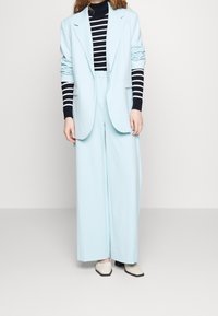 Jasnoniebieska oversize marynarka z szerokimi klapami, zestawiona z granatowo-białym paskowanym golfem i szerokimi spodniami o luźnym kroju. Białe buty na blokowym obcasie z zaostrzonym noskiem.