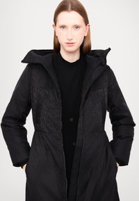 Piumino nero con zip davanti e cappuccio. Presenta un motivo goffrato discreto, polsini a coste e tasche laterali. Tessuto liscio.
