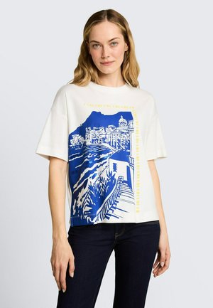 Kobieta o jasnych włosach, ubrana w biały, oversize'owy t-shirt z niebiesko-żółtą grafiką przedstawiającą nadmorskie miasto, w połączeniu z ciemnymi jeansami.