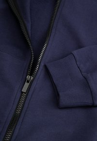 Granatowa bluza na suwak wykonana z miękkiego materiału, z czarnym zamkiem błyskawicznym, ściągaczami na rękawach oraz gładką fakturą. Zbliżenie na widoczne detale.