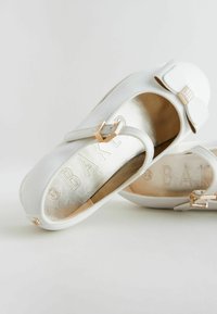 Scarpe bianche dal design flat con un accento a forma di fiocco, realizzate in materiale liscio. Presentano un interno dorato e il marchio 'BAK' impresso sulla suola.