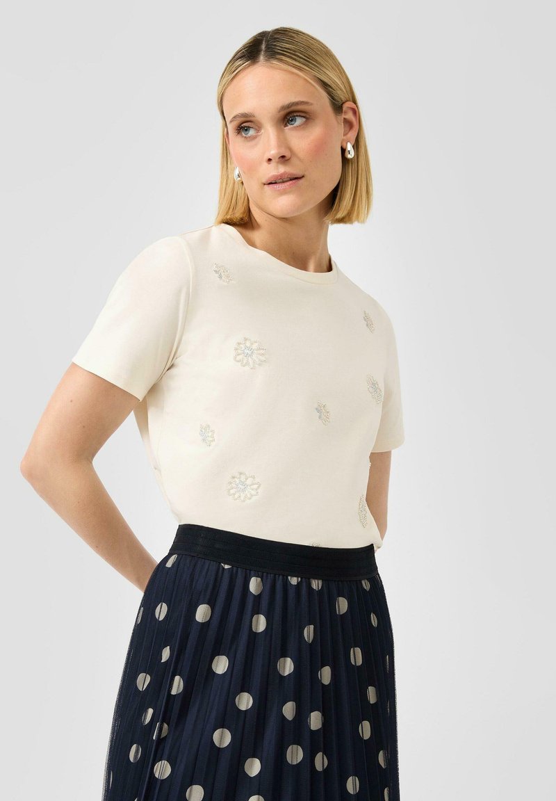 Vrouw met blond bobkapsel, draagt een wit topje met korte mouwen en bloemrijke borduursels, gecombineerd met een marineblauwe geplooide rok met beige polkadotten.