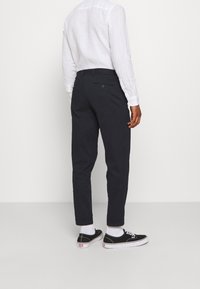 Osoba stojící zády, oblečená v bílém tričku s dlouhým rukávem, černých kalhotách s manžetami, bílých ponožkách a černých nízkých teniskách.