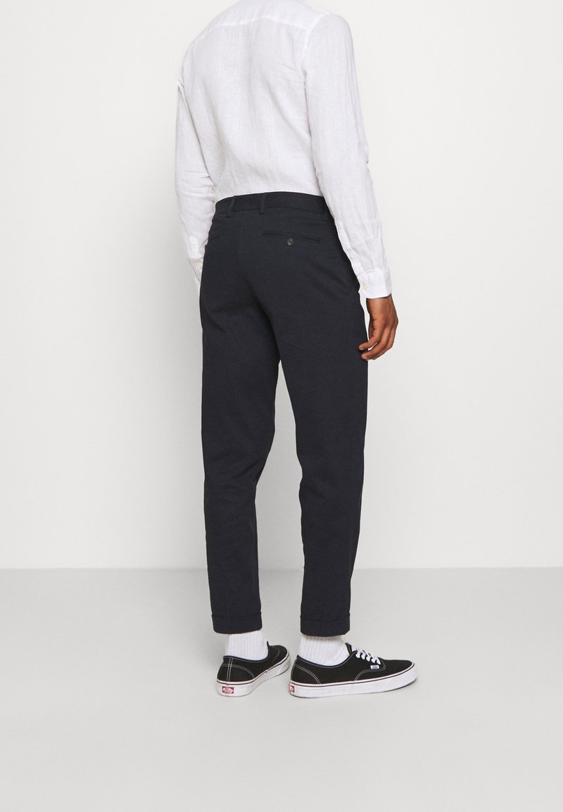 Osoba stojící zády, oblečená v bílém tričku s dlouhým rukávem, černých kalhotách s manžetami, bílých ponožkách a černých nízkých teniskách.