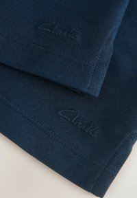Tamsiai mėlynas medvilnės audinys su tekstūruota paviršiumi; turi siuvinėtą logotipą "Clarks" atitinkančia siūle, demonstruodamas švarų dizainą ir kokybišką apdailą.