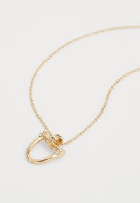 Colar de corrente de ouro com um pendente em forma de U, acentuado por pequenas pedras preciosas transparentes em cada extremidade, sobre um fundo branco.