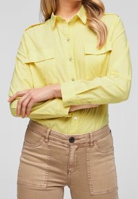 Camicia gialla con bottoni in alto, due tasche sul petto, in tessuto leggero, maniche lunghe e vestibilità rilassata, abbinata a pantaloni skinny beige.