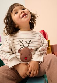 Light-colored striped sweater with a reindeer motif, featuring brown applique and a red nose, paired with ribbed brown pants.