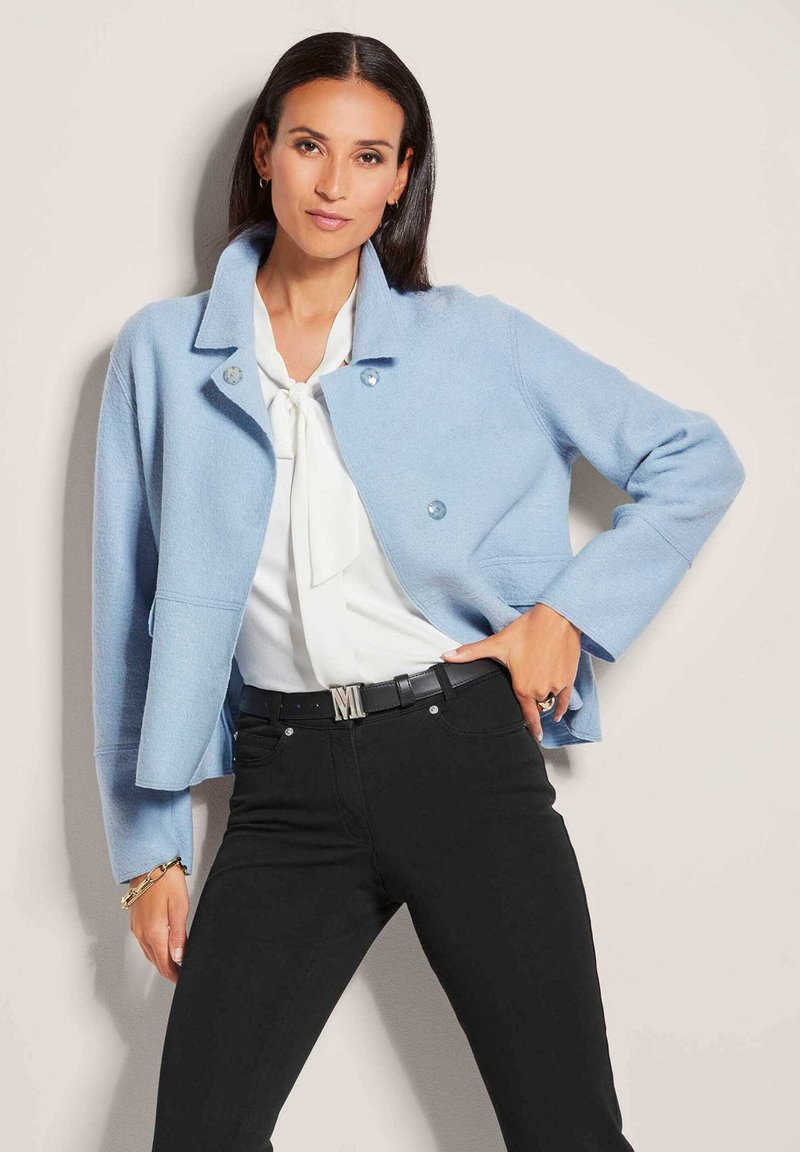 Light blue cropped jacket with a collar, featuring round buttons, worn over a white blouse and black pants. Contains subtle texture and tailored fit.